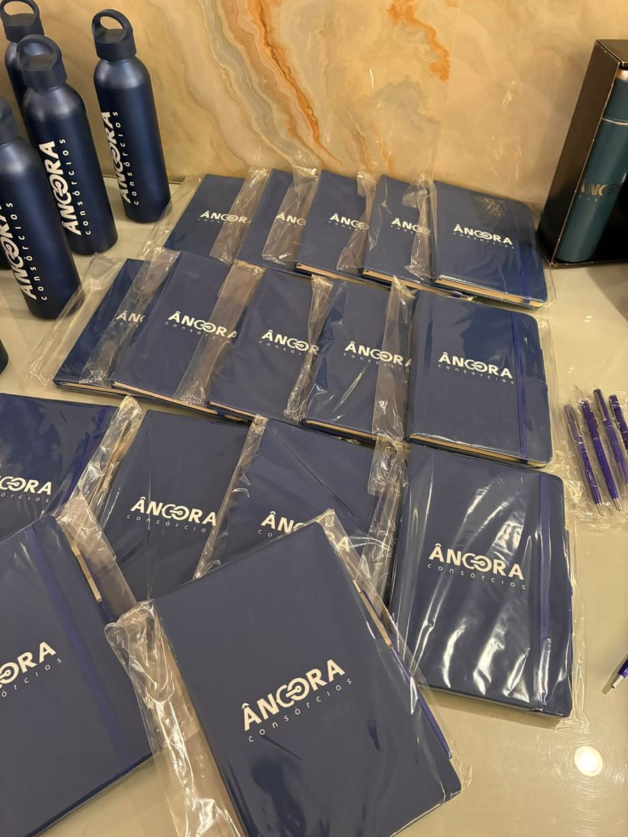 Cadernos azuis personalizados com a logo de uma empresa de consórcios, ao lado de canetas e garrafas da mesma marca